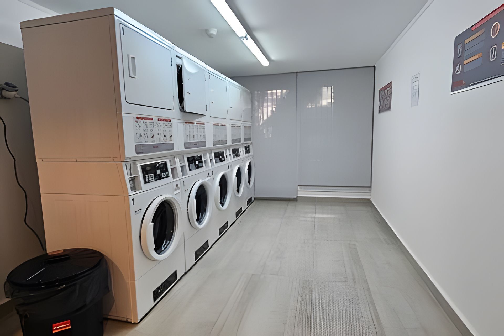 Un cuarto de lavandería con varias lavadoras, un cubo de basura y un panel de control en la pared.
