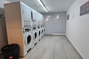 A laundry room with multiple washing machines, a trash bin, and a wall-mounted control panel.