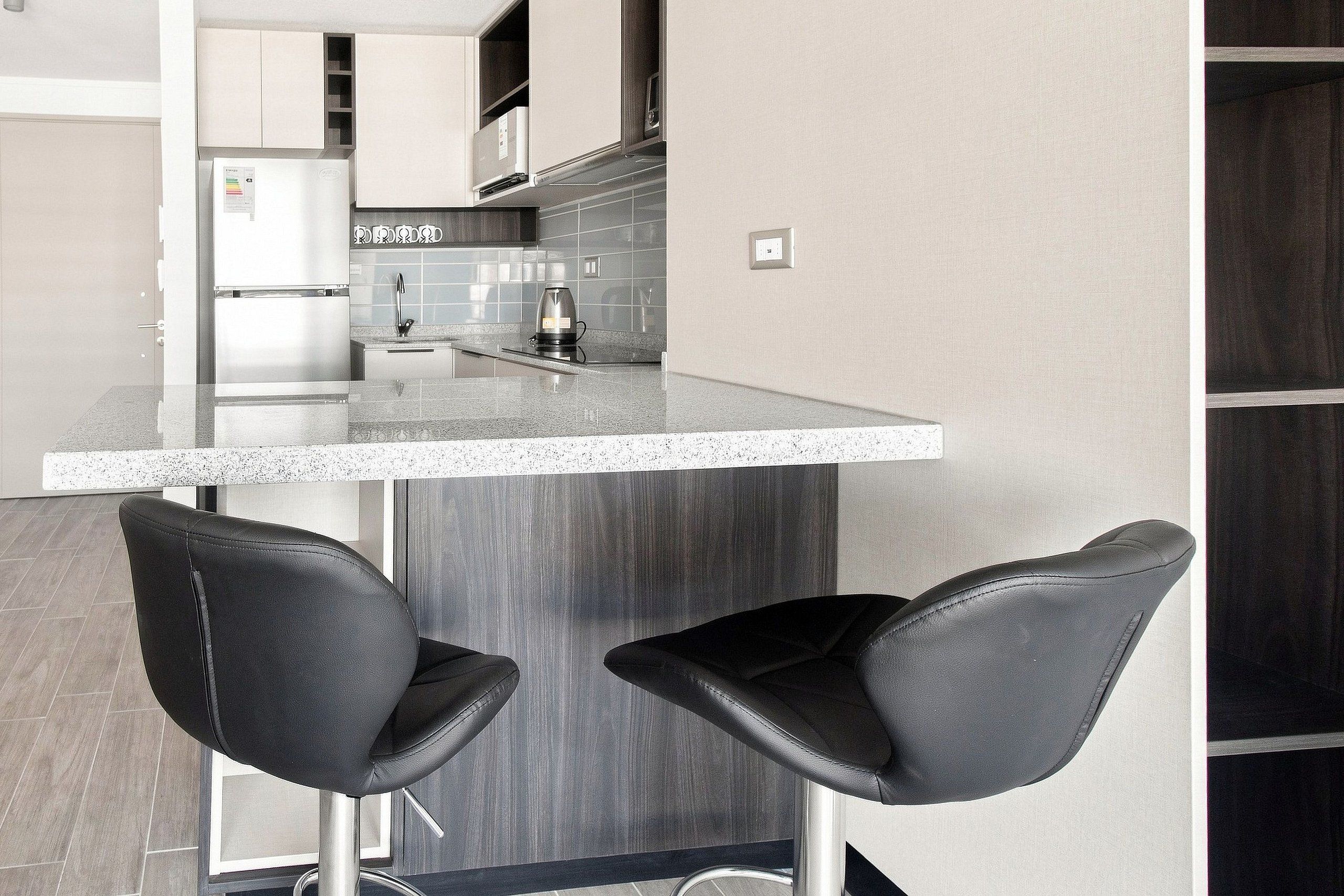 Una cocina moderna con barra, dos taburetes negros y un refrigerador de acero inoxidable.