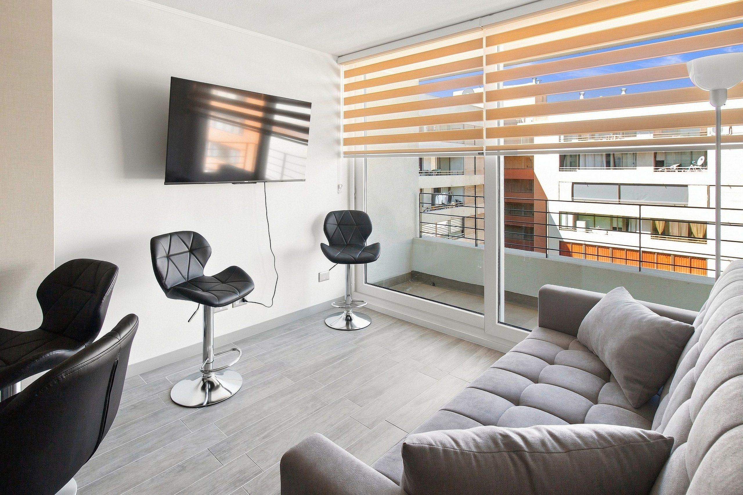 Una sala de estar moderna con un sofá gris, dos sillas negras, un televisor montado en la pared y un balcón con vista a un edificio.