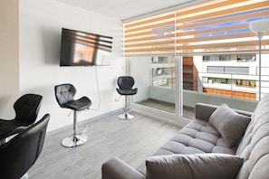 A modern living room with a grey sofa, two black chairs, a TV mounted on the wall, and a balcony with a view of a building.