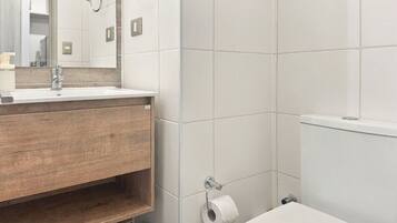 Un baño con un inodoro blanco, un tocador de madera, un espejo y un rollo de papel higiénico en la pared.