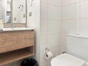 Een badkamer met een wit toilet, een houten wastafelmeubel, een spiegel en een rol toiletpapier aan de muur.