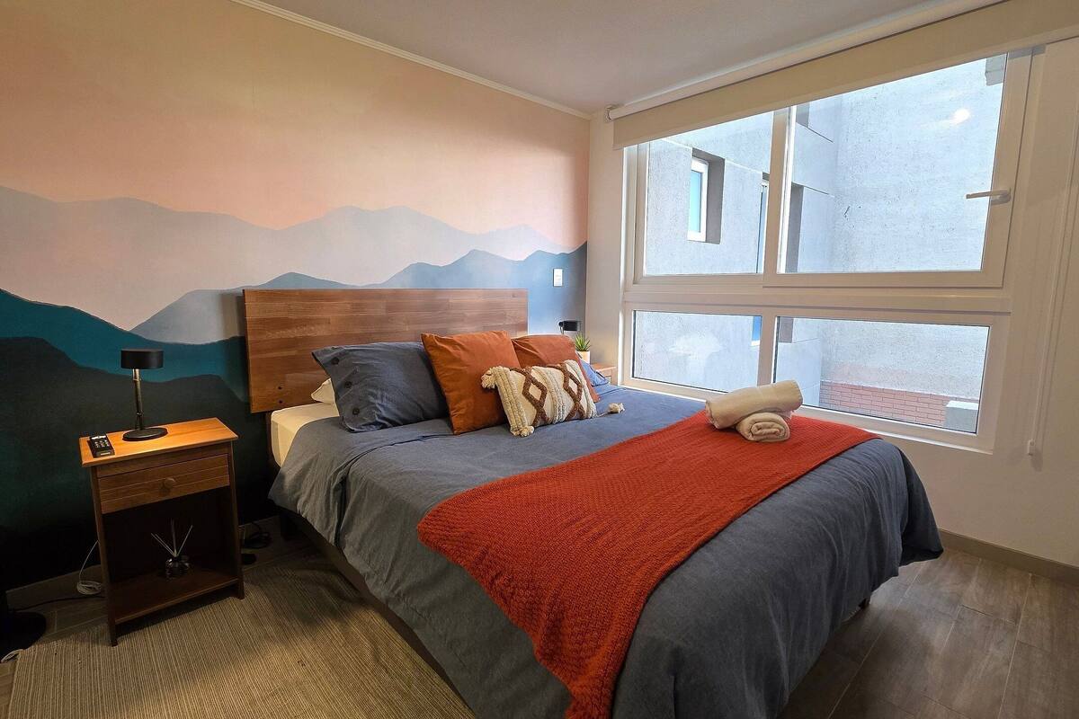 Een slaapkamer met een bed, nachtkastje, lamp en uitzicht buiten door een raam.