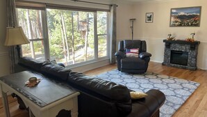 A living room with a leather sofa, a small table with a lamp, a fireplace, and a large window with a view of trees. - Spacious 4-bed house in the woods of beautiful Monument by the Air Force Academy (Monument)