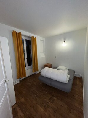 A bedroom with a bed, a nightstand, a window with curtains, and a wall-mounted light.