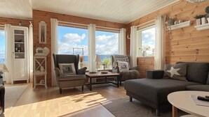 Una sala de estar acogedora con un sofá, sillones y una mesa de centro. Hay ventanas amplias con cortinas, techo de madera y vistas al exterior.