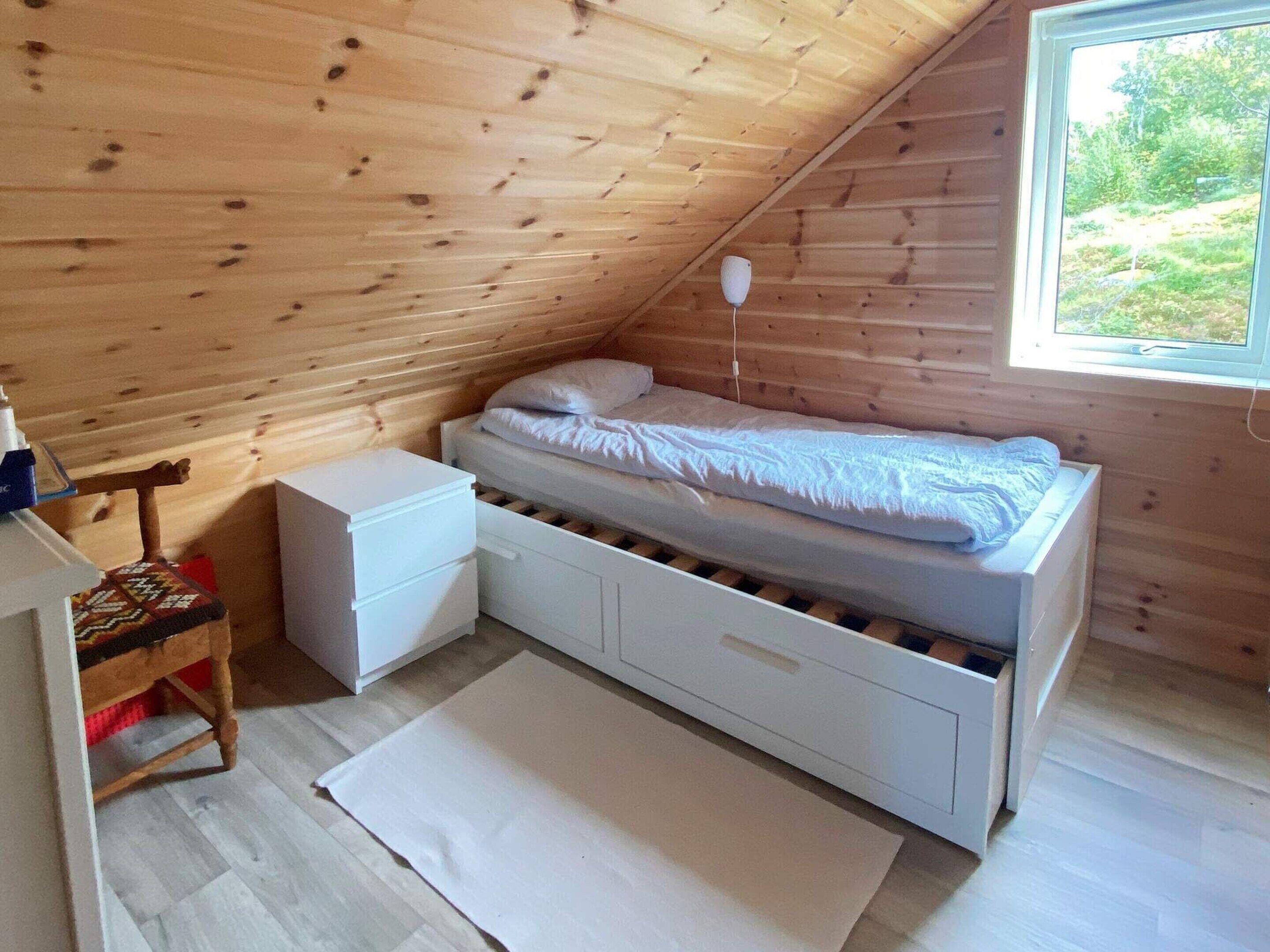 Un dormitorio pequeño y acogedor con techo inclinado, una cama individual, una mesita de noche, un aparador y una ventana con vistas a la vegetación.