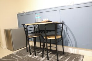 Ein kleiner, schwarzer Nachttisch aus Metall mit drei Ablagen und einem Hocker, platziert in einer Ecke, im Hintergrund ein Kühlschrank und eine blaue Wand.