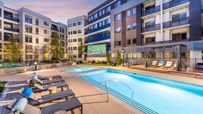 A modern hotel with a swimming pool, lounge chairs, and a large screen displaying a sports event.