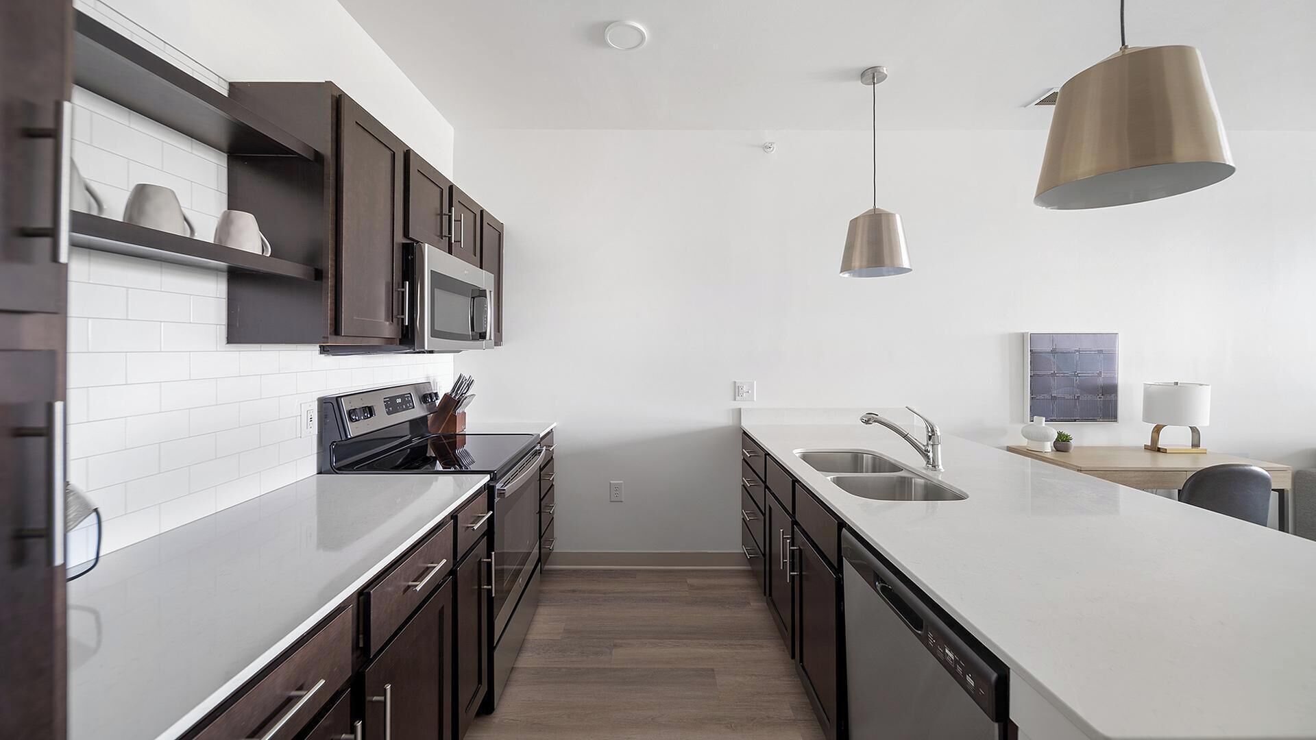 A modern kitchen with dark cabinetry, white countertops, a built-in oven, a microwave, and a large island with a sink. There are pendant lights above the island.