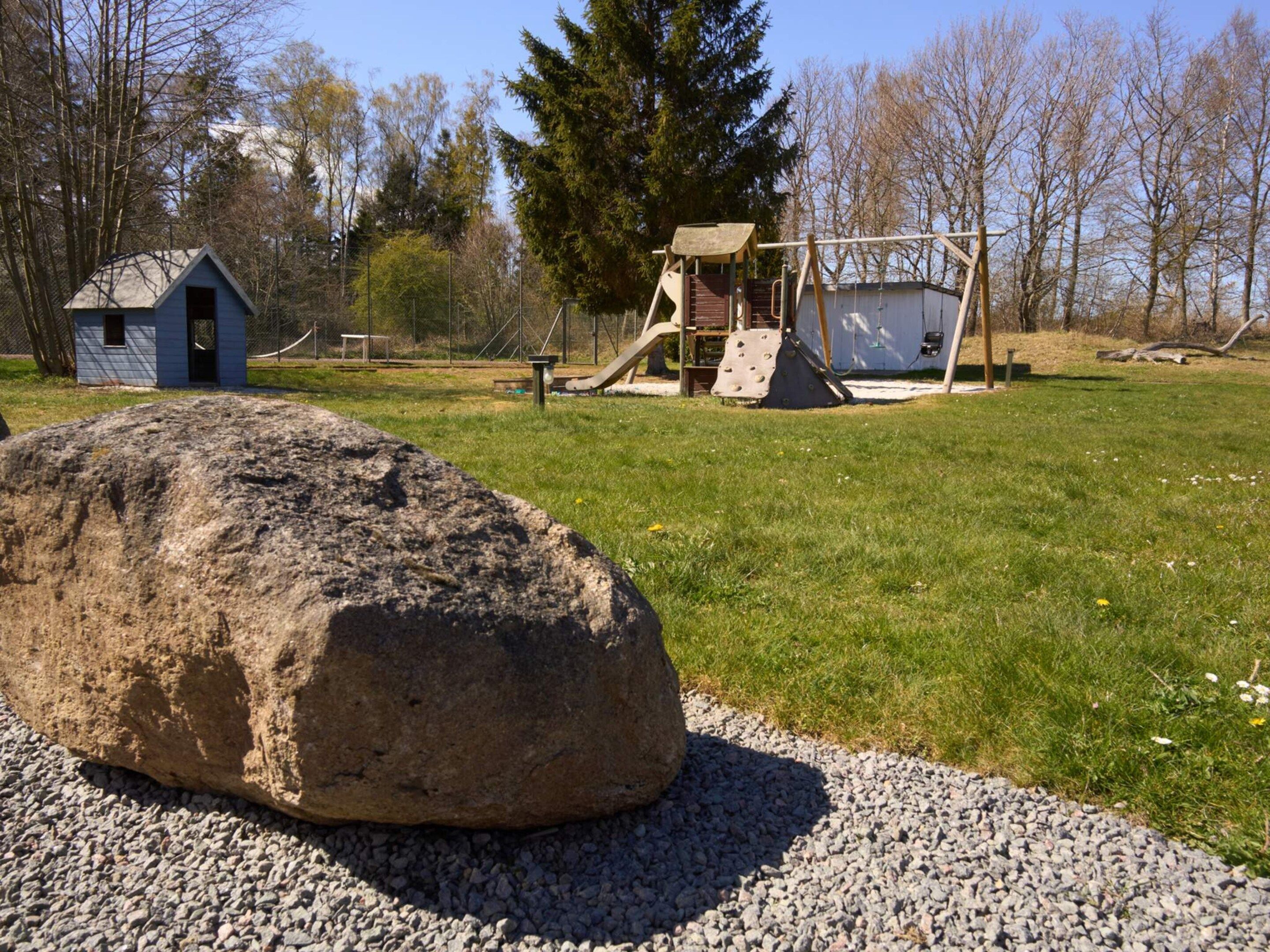 A playground with a swing set and climbing structures, a small shed, and a large rock in a grassy area with trees in the background.