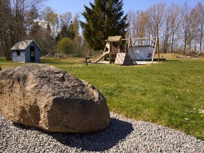 Ein Spielplatz mit Schaukeln, Klettergerüsten, einer kleinen Gartenhütte und einem großen Felsen in einer grünen Wiese, im Hintergrund Bäume.