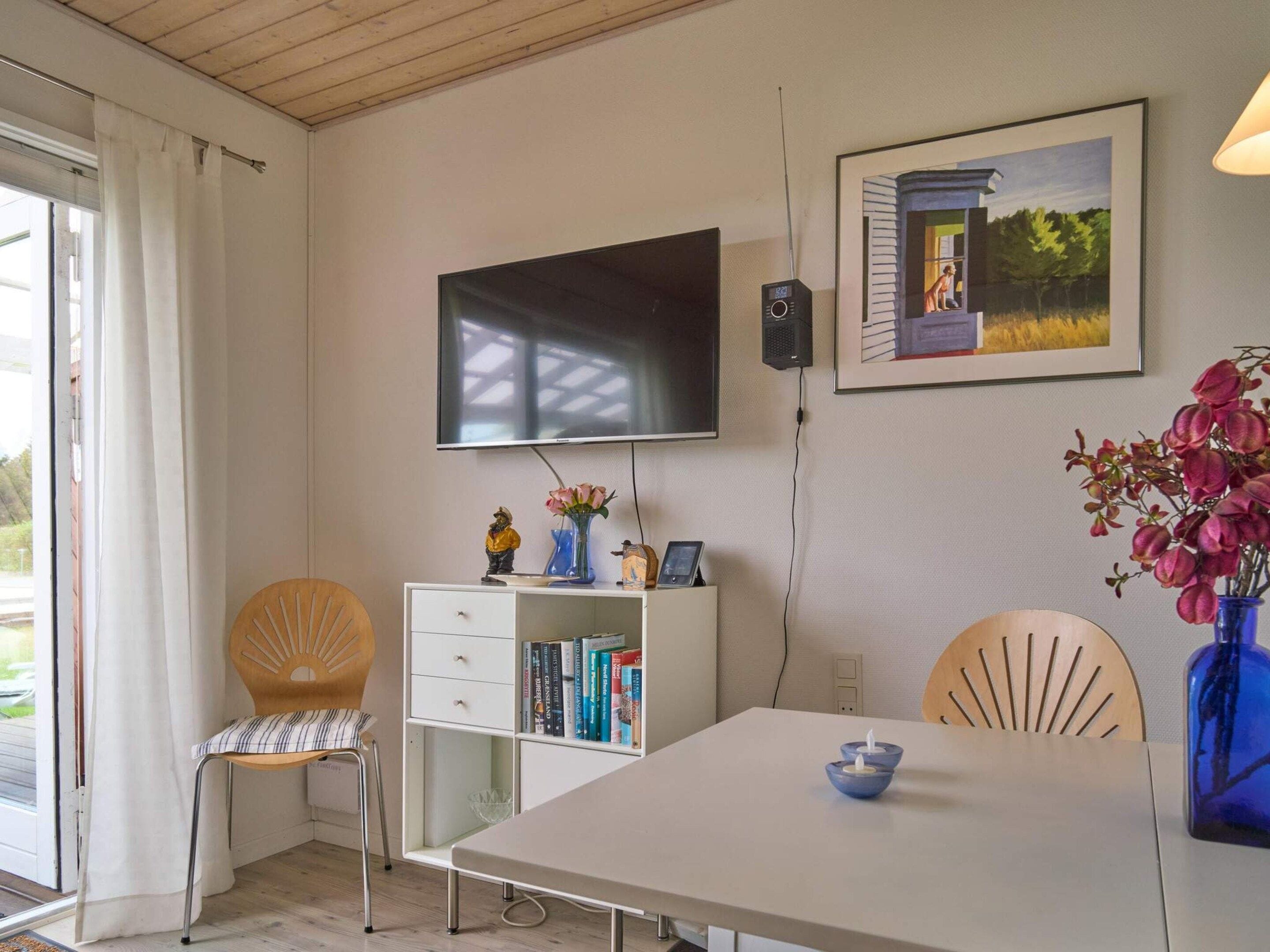 Una habitación con una televisión de pantalla plana montada en la pared, una estantería con libros, una mesa de comedor con una vela azul y una silla con un diseño tipo sol.
