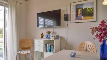 Una habitación con una televisión de pantalla plana montada en la pared, una estantería con libros, una mesa de comedor con una vela azul y una silla con un diseño tipo sol.