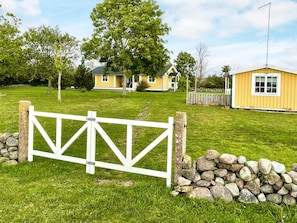 A gated entrance to a residential area with a grassy lawn, a stone wall, and a yellow building with a gabled roof.