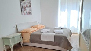 A neatly made bed with a grey and peach bedspread, a small bedside table with a greenish hue, and a large window with sheer curtains.