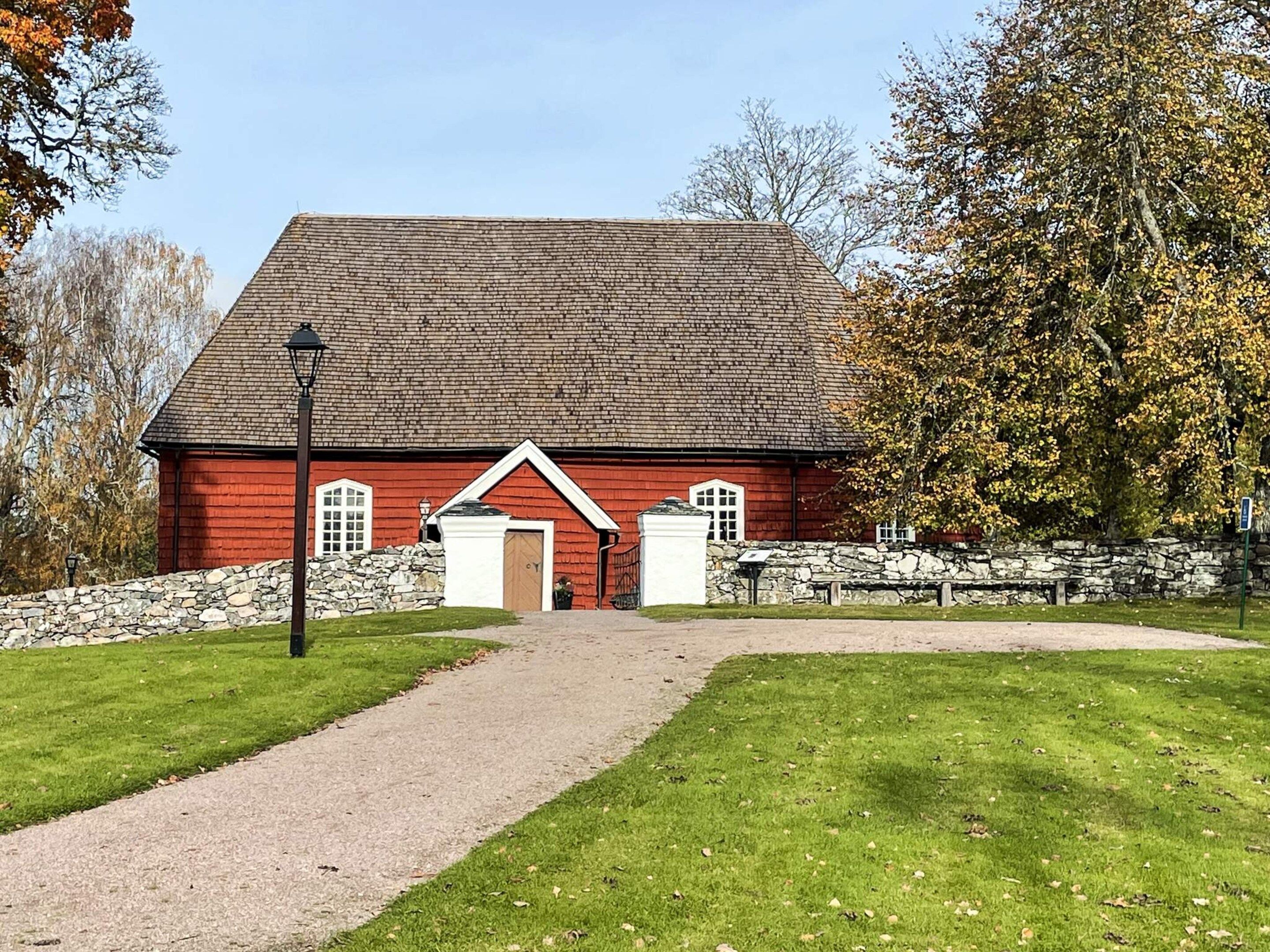 A red building with a stone wall, a paved path leading to the entrance, and a bench area to the right.