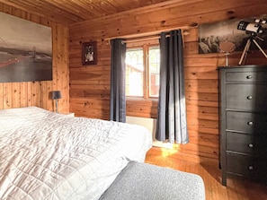 Une chambre dans un chalet en bois, avec un lit, une commode et un télescope monté sur un trépied.