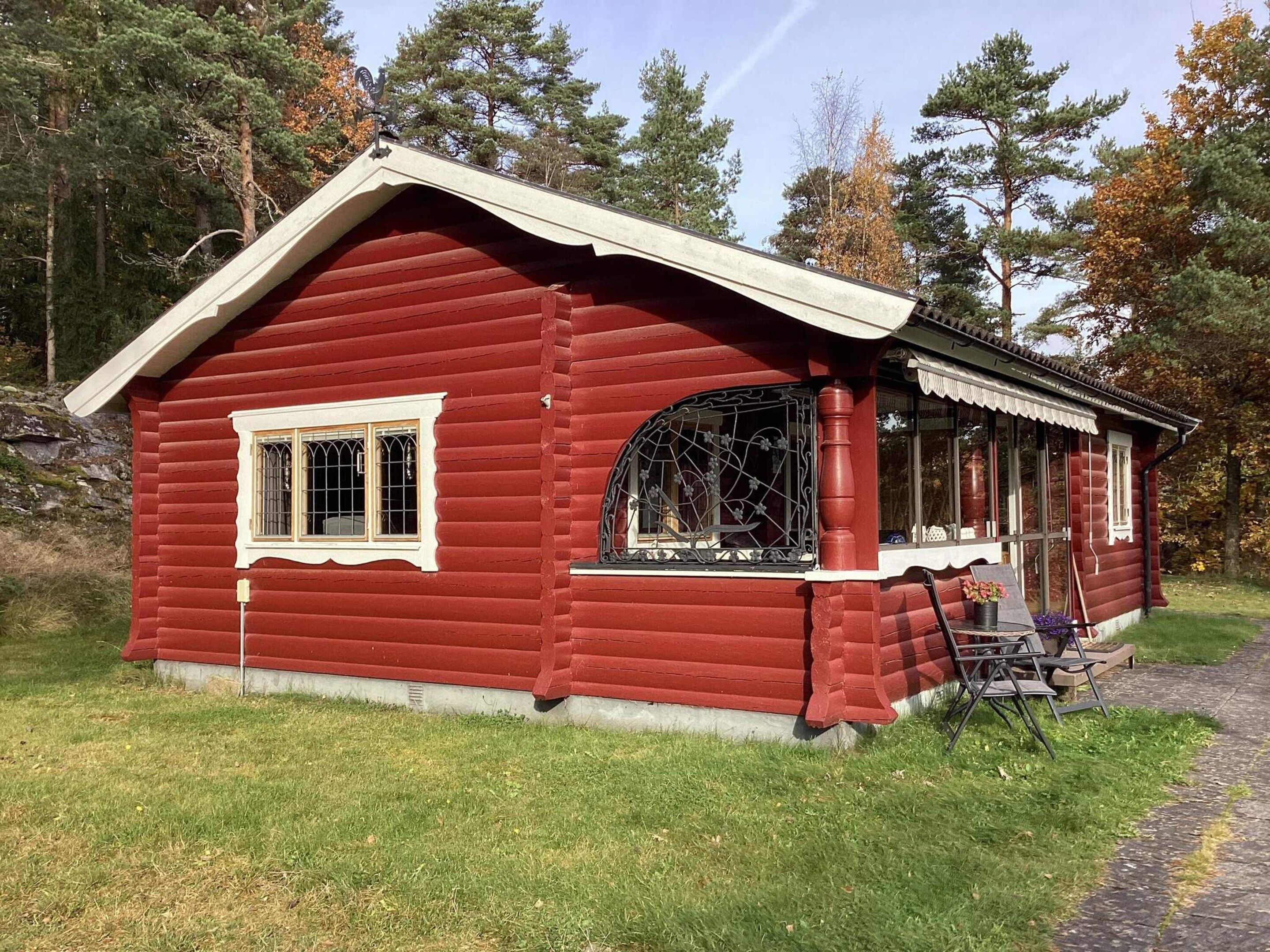 A red log cabin with a porch, a window with decorative iron bars, and a small garden area with a chair and plants.