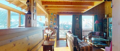 Uma cabana de madeira com área de jantar, sofá e vista para montanhas nevadas.