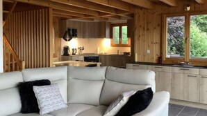 A modern log cabin interior with a living area featuring a white leather sofa and black cushions, a kitchen with wooden cabinets and a window, and a staircase leading to an upper level. - Chalet in the Heart of Valley With Balcony (Nendaz)