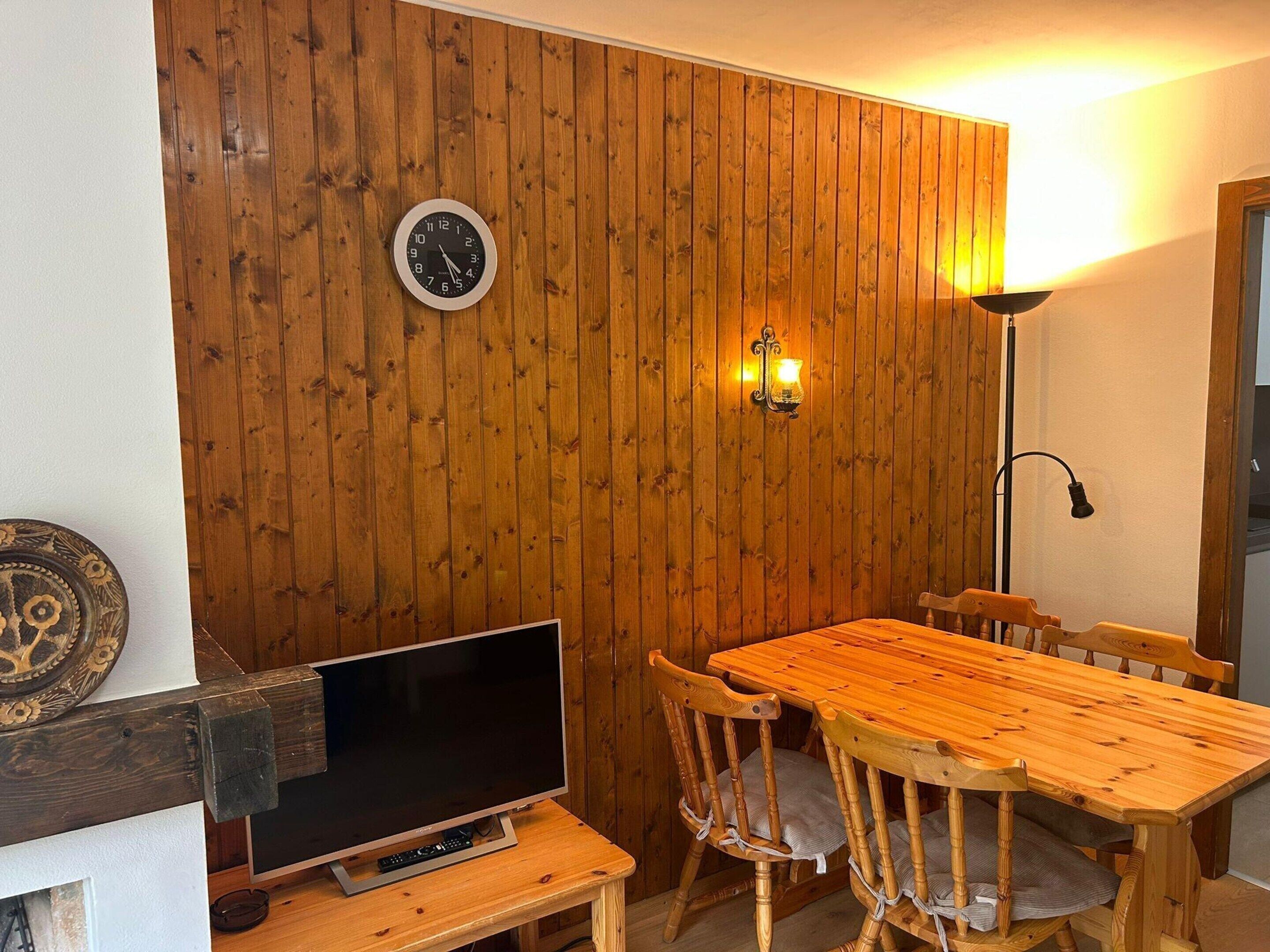 A wooden-paneled room with a wooden dining table, chairs, a television on a stand, a wall clock, and a wall-mounted light fixture.