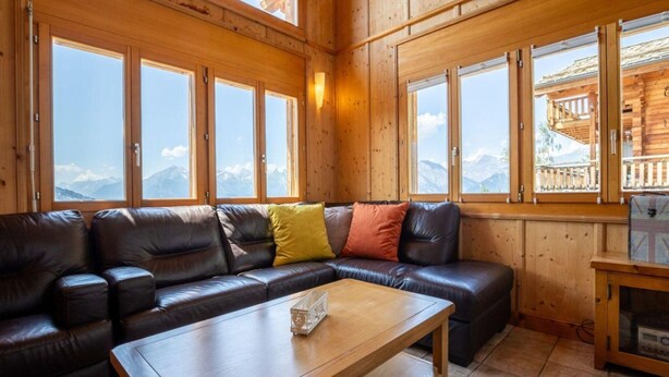 Interno di una baita in legno con un divano in pelle, un tavolino in legno e ampie finestre con vista sulle montagne.