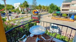 Un balcón con sillas de mimbre y una mesa, con vista a una calle donde se ve un cartel de hotel, palmeras y coches aparcados.