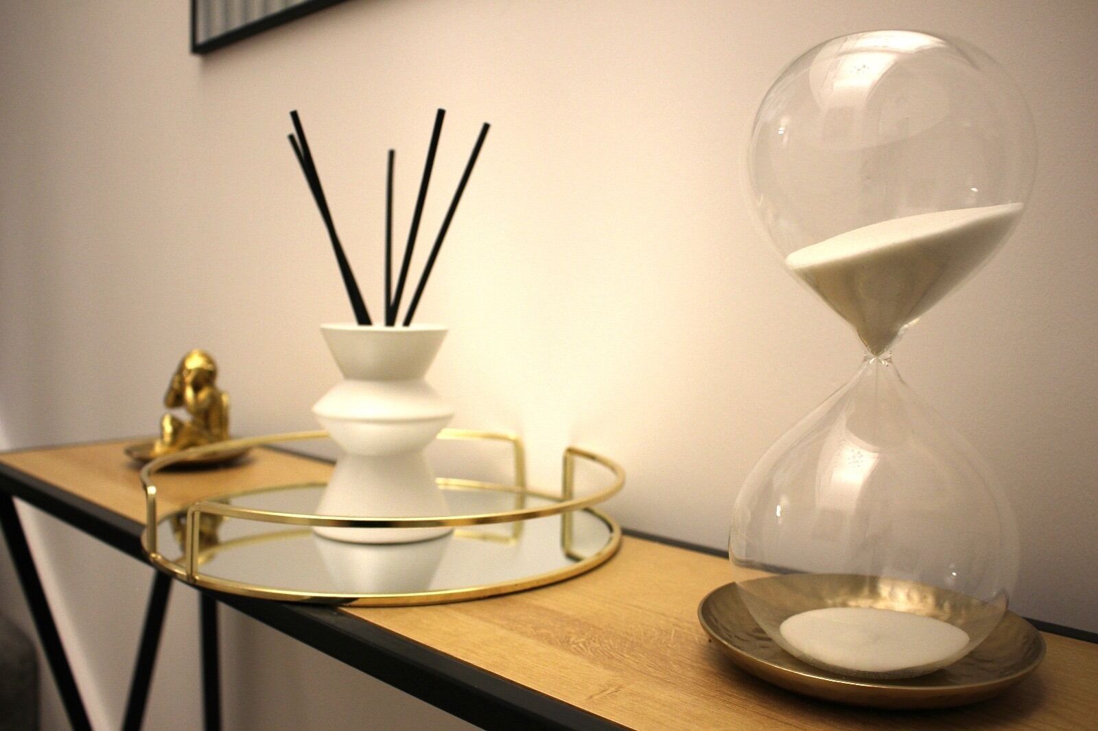 A table with a white vase containing black sticks, a golden hourglass, and a golden tray.