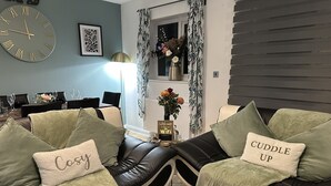 A modern living room with a black leather sofa, two cushions with text, a round wall clock, and a dining area in the background. - Bramblemead 5 bed House in Manchester Leigh (Leigh)