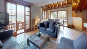 A spacious living room with a sofa, a coffee table, and a TV. There are large windows and a balcony with a wooden railing. - Comfortable Apartment With Sauna (Veysonnaz)