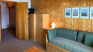 A compact room with a wooden wardrobe, a green sofa with patterned cushions, a small table, and a wall-mounted lamp. - Luxury Studio 4 Pers (Veysonnaz)