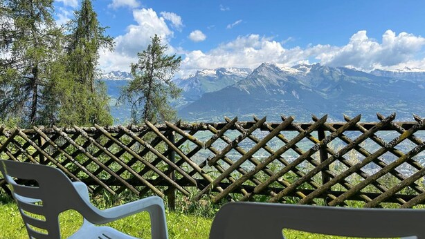 A view from a balcony with a wooden lattice fence, two white plastic chairs, and a backdrop of snow-capped mountains under a clear blue sky. - Luxury Studio 4 Pers (Veysonnaz)