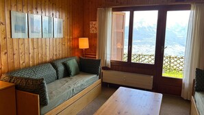 A wooden cabin room with a sofa, a table, and a window with a view of mountains. - Luxury Studio 4 Pers (Veysonnaz)