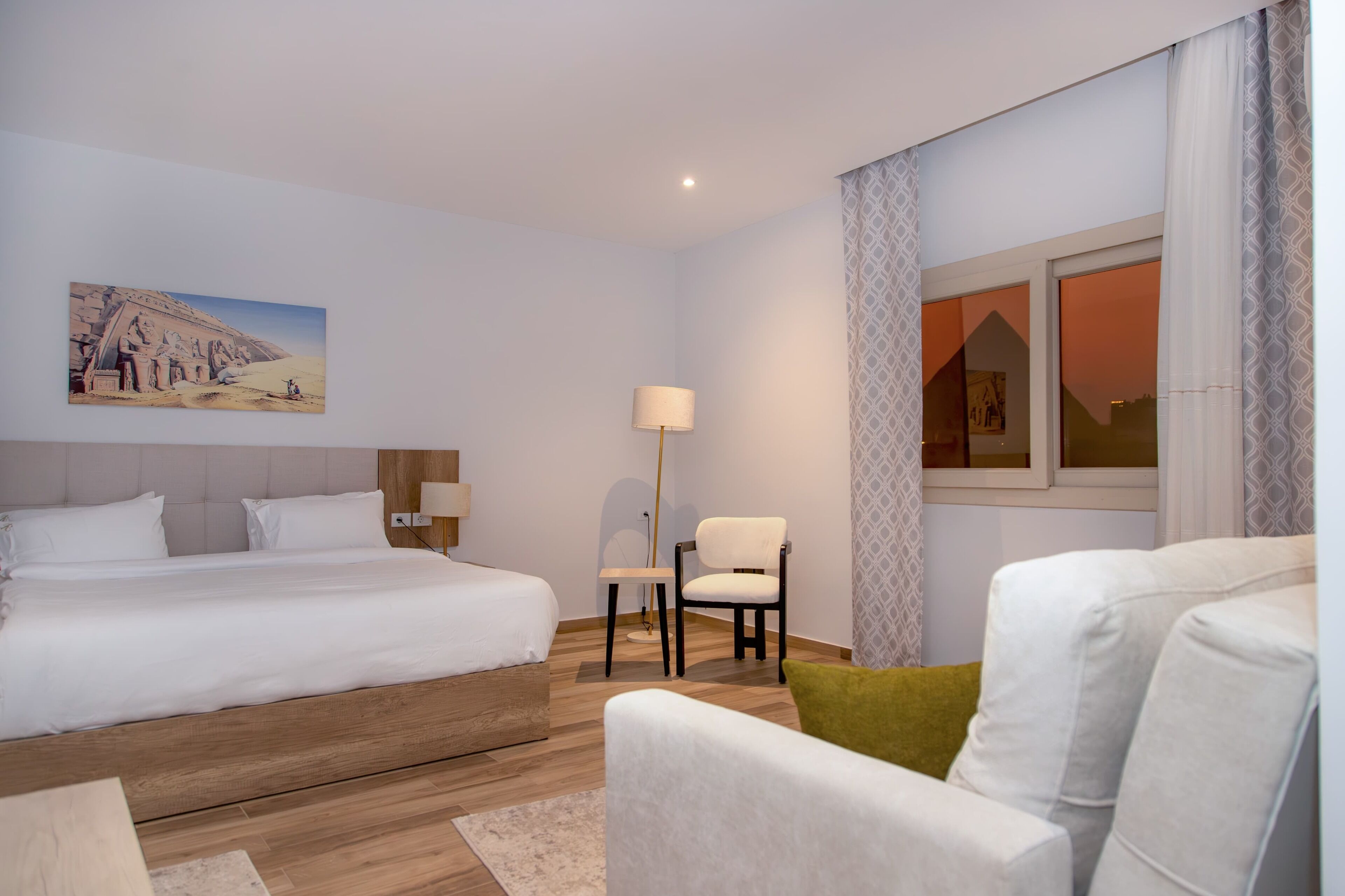Habitación de hotel con una cama grande, una cama más pequeña, una silla, una lámpara y un cuadro en la pared.