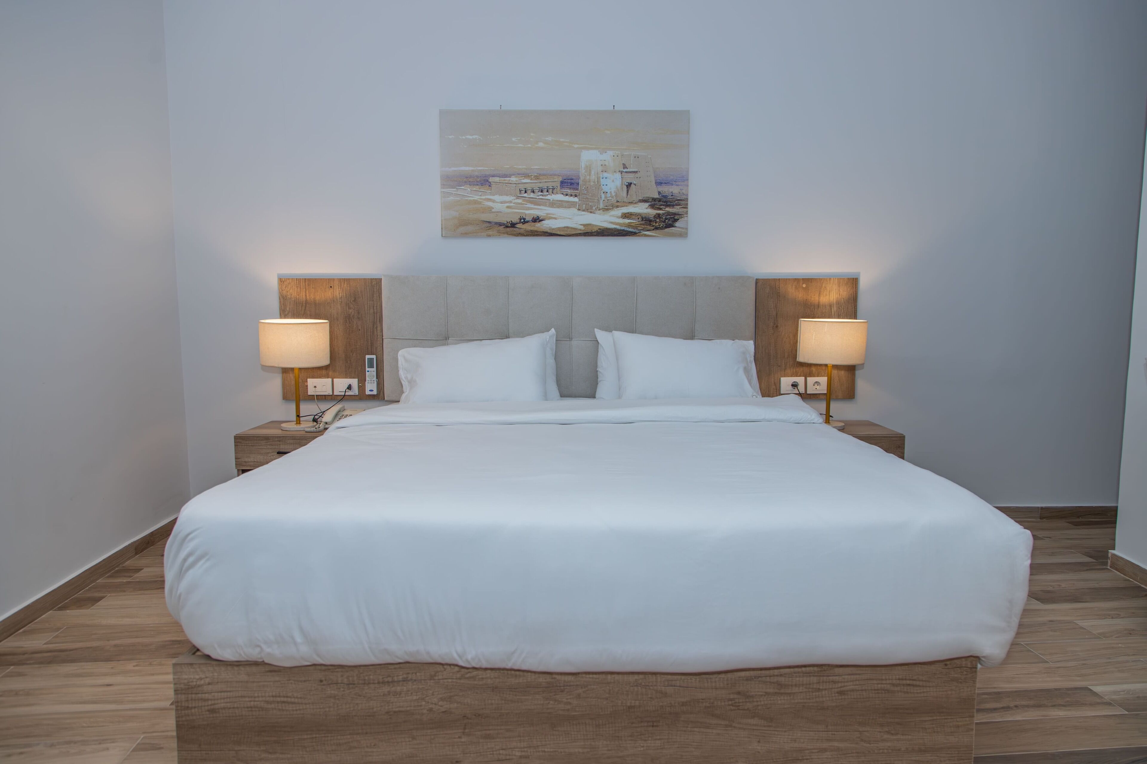 A neatly made bed with white linens, two bedside lamps, and a painting of a castle on the wall.
