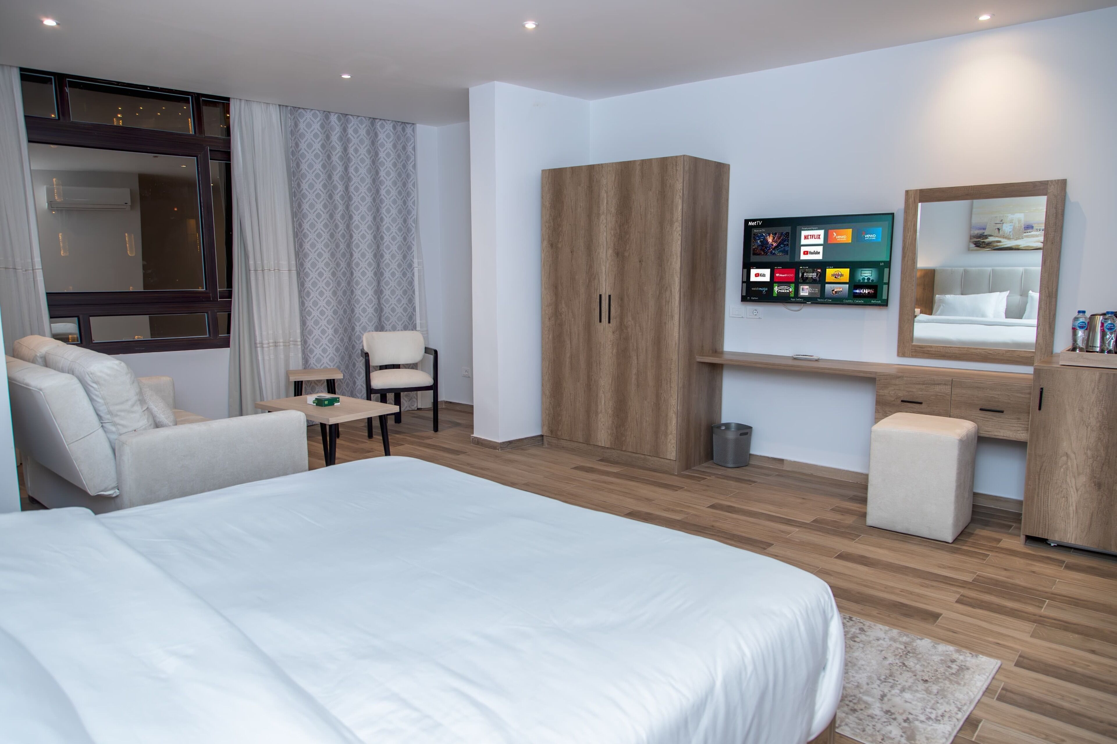A modern hotel room with a large bed, a sofa, a desk, a wardrobe, and a TV displaying streaming services.