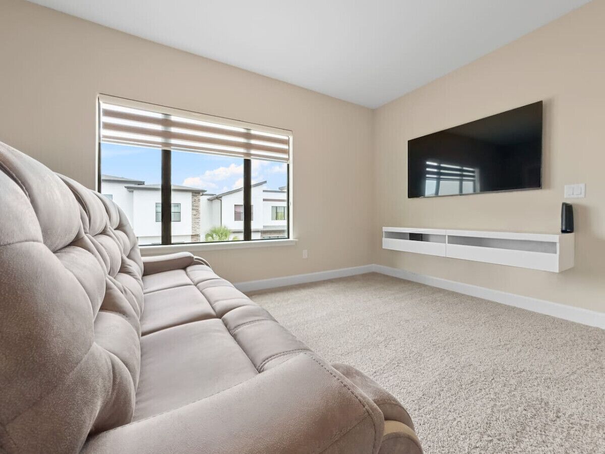 Un salon avec un canapé beige, une grande fenêtre avec des stores, un téléviseur à écran plat fixé au mur et un meuble de rangement blanc.