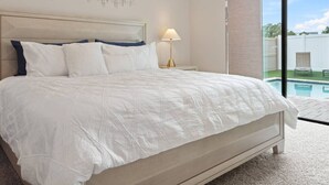 A bedroom with a large bed, a nightstand, a lamp, and a view of a swimming pool through a sliding glass door. - Dolce Far Niente - Villa Domani (Davenport)
