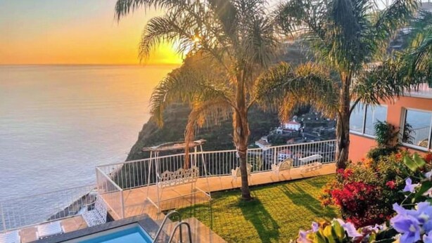 A pool area with a view of the ocean at sunset, surrounded by palm trees and a small garden. - Cliff House (Lombo do Doutor)