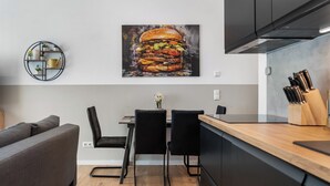 Cozinha moderna com área de refeições, com um quadro grande pendurado na parede retratando um hambúrguer, uma prateleira de metal redonda e bancada de madeira com utensílios de cozinha.