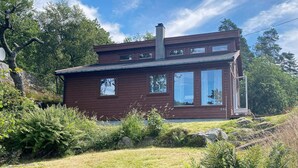 A two-story wooden cabin with a chimney, large windows, and a grassy yard with rocks and ferns. - 7 Person Holiday Home in Lyngdal (Lyngdal)