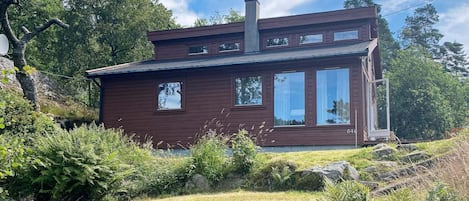 A two-story wooden cabin with a chimney, large windows, and a grassy yard with rocks and ferns.