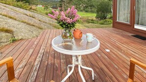 A wooden deck with a round glass table, a vase with pink flowers, a teapot, and a mug. - 7 Person Holiday Home in Lyngdal (Lyngdal)
