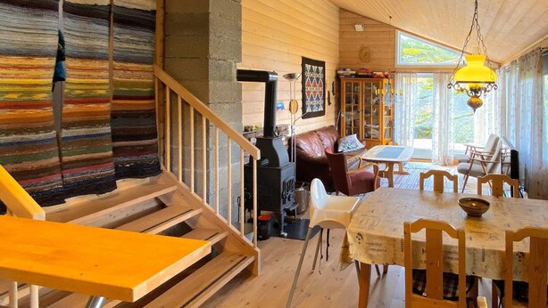 A cozy cabin interior with a wooden staircase, a dining area with a table and chairs, a fireplace, and a large window providing natural light. - 7 Person Holiday Home in Lyngdal (Lyngdal)