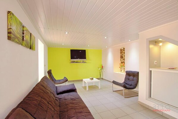 A modern living room with a green accent wall, a flat-screen TV, a sofa, two armchairs, and a coffee table.