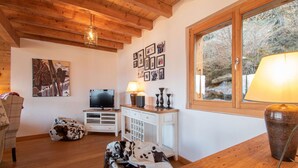 Ein gemütliches Wohnzimmer mit Holzboden, einem Fernseher, einem weißen Schrank, einem Fenster mit Blick auf Bäume und einem Sitzsack mit Kuhmuster.