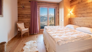 Ein Schlafzimmer mit Holzboden, einem Bett mit weißer Bettwäsche, einem Korbstuhl, einem Fenster mit Vorhängen und Blick auf die Berge.