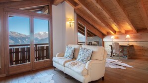 Ein gemütliches Zimmer mit Holzbalkendecke, einer Couch mit gemusterten Kissen, einem Esstisch mit Stühlen und Blick auf Berge durch große Fenster.